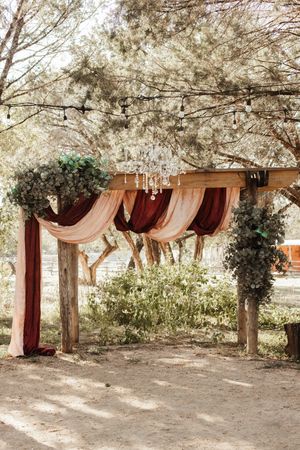Rustic outdoor wedding arch with burgundy and blush draping, crystal chandelier, and lush greenery decor.