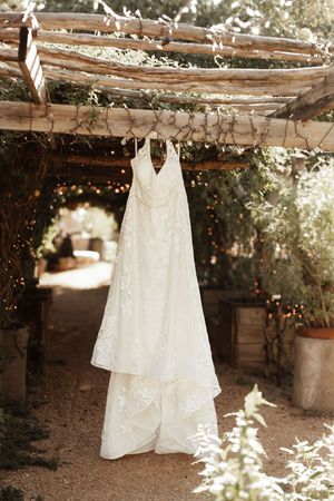 Elegant white lace wedding dress hanging on rustic wooden pergola surrounded by greenery and fairy lights.