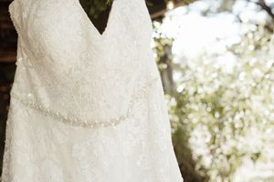 Elegant white lace wedding dress with beaded belt hanging outdoors with soft bokeh greenery background.