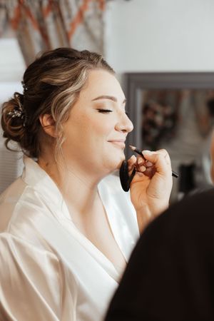 Bride in white robe getting makeup applied by artist, smiling with elegant updo hairstyle.
