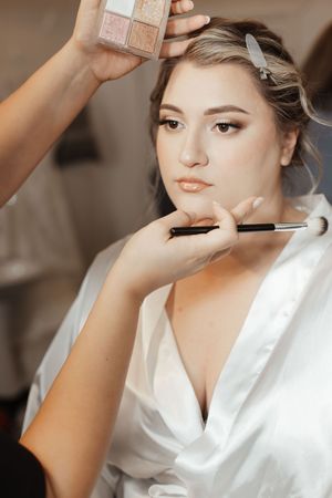 A bride in a white satin robe gets professional makeup applied with a brush while a makeup palette is held nearby.