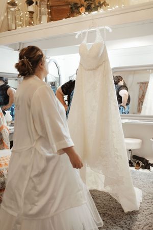 Bride in white robe admiring her elegant lace wedding gown hanging in a bridal suite before the ceremony.