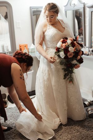 Bride in lace wedding gown holding bouquet while bridesmaid adjusts her dress train before the ceremony.