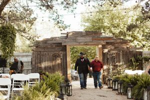 Two men walk hand-in-hand through a rustic wooden archway at an outdoor wedding ceremony with white chairs and lanterns.