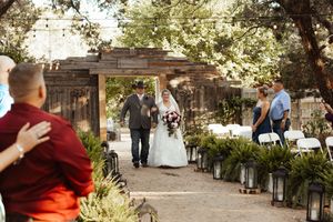 Bride and groom walk down outdoor wedding aisle lined with lanterns and ferns under rustic wooden arbor.