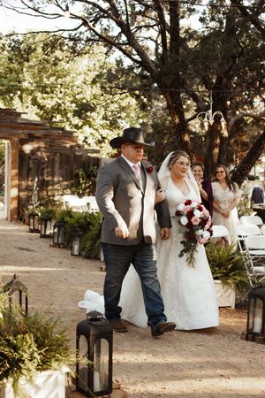 Bride in white lace gown walks down outdoor aisle with man in cowboy hat and gray suit at rustic wedding ceremony.
