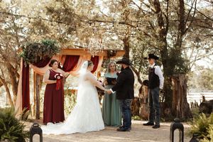 Outdoor wedding ceremony under trees with bride, groom, officiant, and wedding party in burgundy and sage green attire.