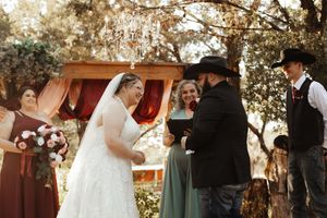 Outdoor wedding ceremony under oak trees with bride in white gown and veil exchanging vows with groom in cowboy hat.