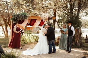 Outdoor wedding ceremony with bride and groom kissing at rustic floral arch, surrounded by wedding party in burgundy and sage green.