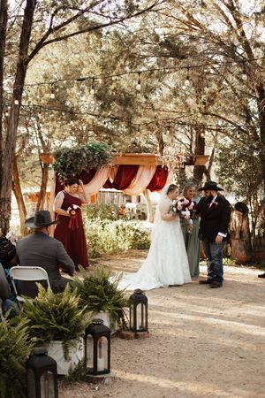 Outdoor rustic wedding ceremony under tree canopy with bride in white gown, floral arch, lanterns, and guests in cowboy hats.