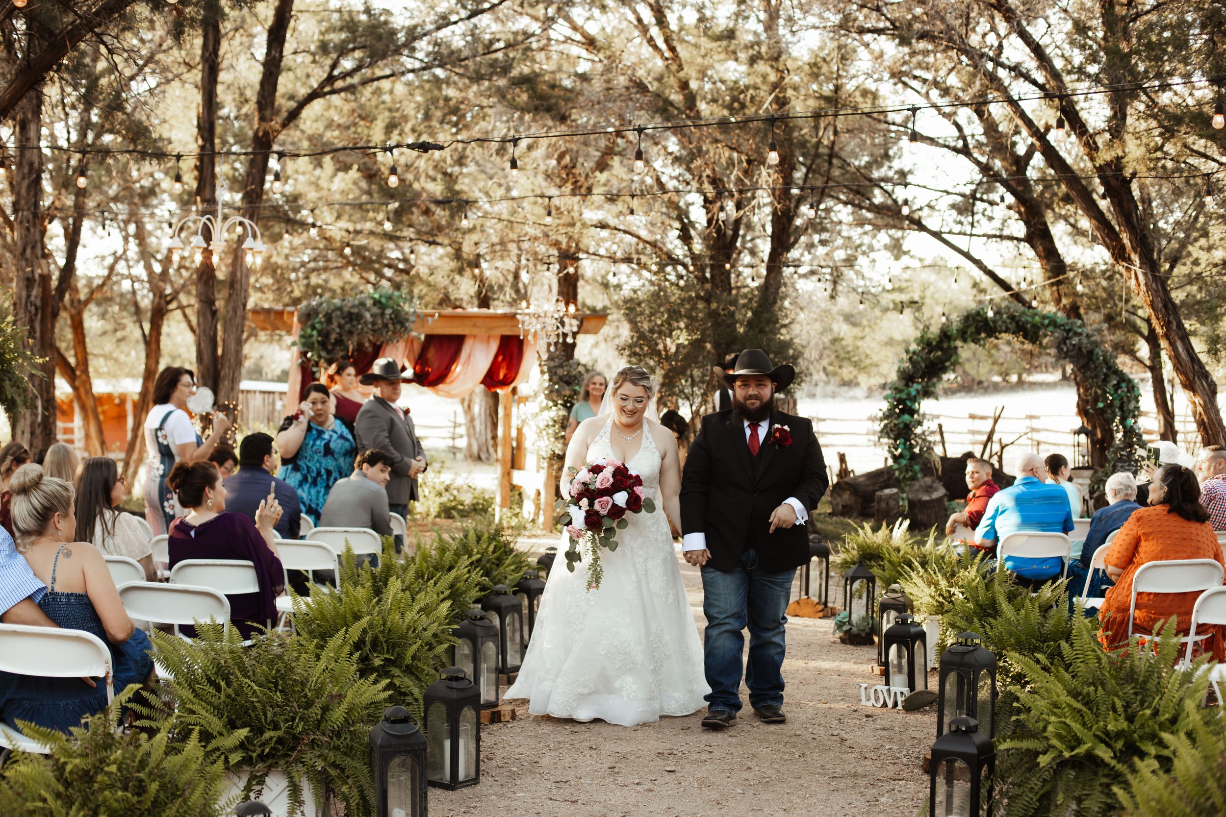 Bride and groom walk down outdoor wedding aisle surrounded by guests, ferns, and lanterns beneath shaded trees.