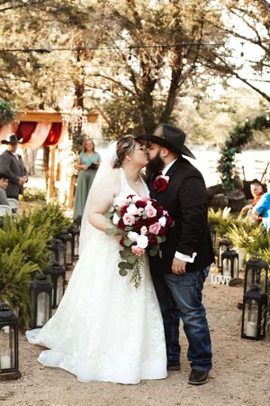 Bride and groom share first kiss at outdoor rustic wedding ceremony, bride holding red and white bouquet, groom in cowboy hat.