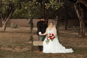 Bride in white gown and groom in black suit pose on a rustic wooden swing surrounded by trees.