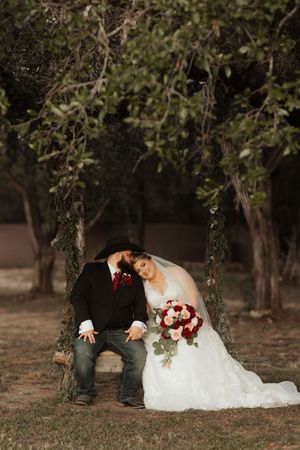 Bride and groom sitting on a wooden swing under trees, bride holding red and white bouquet in wedding attire.