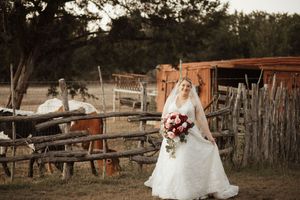 Bride in white gown and veil holding bouquet poses by rustic wooden fence on farm with horses at golden hour.