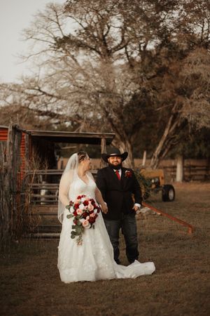 Bride and groom in western attire pose on a rustic farm, she holds red and pink bouquet, he wears black cowboy hat.