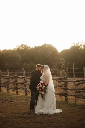 A bride and groom share a romantic kiss at golden hour beside a rustic wooden fence at an outdoor farm wedding.