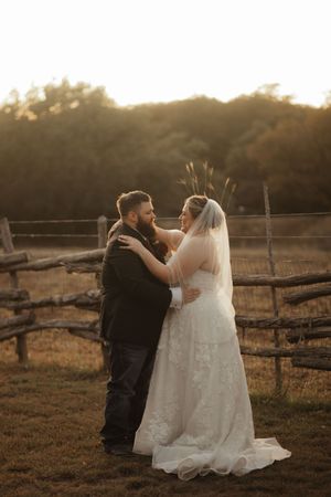 Bride and groom embrace at golden hour beside a rustic wooden fence in an open field, wearing wedding gown with veil.