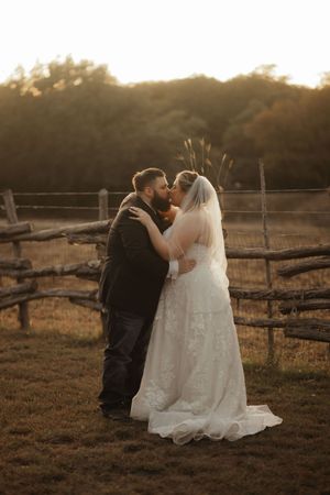 Bride and groom share a romantic kiss at golden hour by a rustic wooden fence in an outdoor wedding setting.