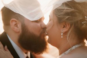 Bride and groom sharing a romantic kiss under a flowing veil in warm golden sunlight on their wedding day.