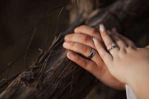 Two hands resting on driftwood, each wearing wedding rings, symbolizing love and commitment.