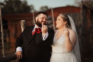 A bride and groom share a joyful moment outdoors, with the bride in a lace gown and veil and groom in a dark suit with red tie.