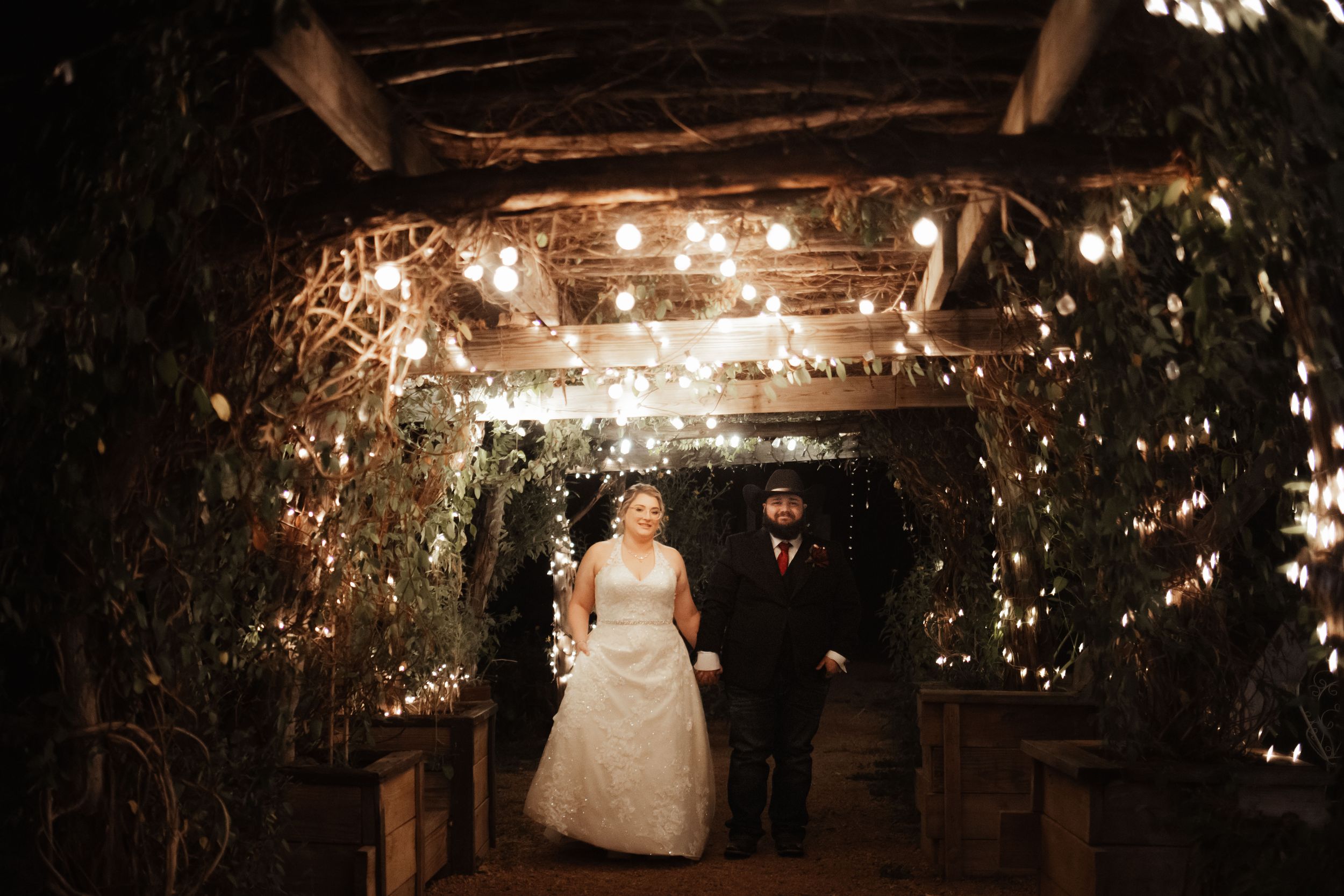Bride and groom walk hand in hand through a romantic, fairy light-adorned garden tunnel at their evening wedding reception.