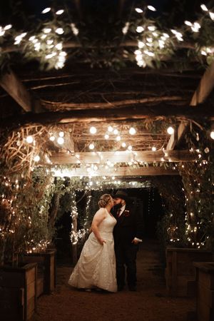 Bride and groom sharing a romantic kiss under glowing string lights and lush greenery at an enchanting outdoor wedding venue.