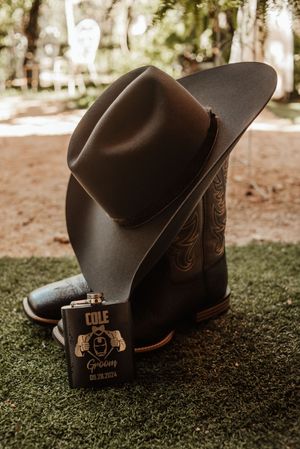Brown cowboy hat and boots with a personalized groom flask reading Cole, displayed on green grass for a rustic wedding.