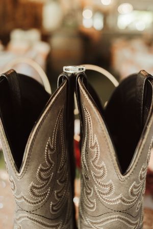 Wedding rings resting between two gray cowboy boots with intricate stitching at a rustic country wedding venue.