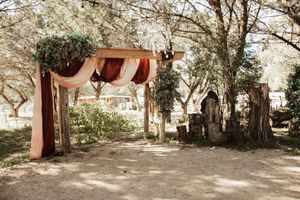 Rustic outdoor wedding arch adorned with draped fabric and greenery among trees in a natural garden setting.