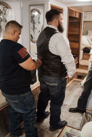 Men helping a groom in a black vest and jeans get dressed in a rustic room before a wedding ceremony.