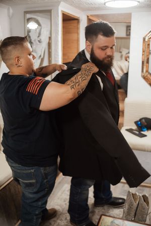 Tattooed man helping bearded groom put on black suit jacket while getting ready indoors.