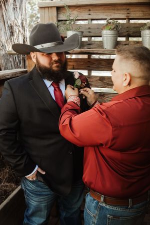 A bearded groom in a black suit and cowboy hat gets his boutonniere pinned by a man in a red shirt at a rustic wedding.