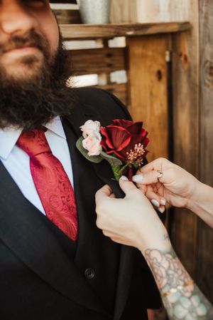 Tattooed hand pins red rose boutonniere on bearded groom in black suit with red paisley tie.