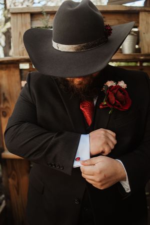 Bearded groom in black suit, red tie, boutonniere, and cowboy hat adjusting cufflinks at rustic wedding.