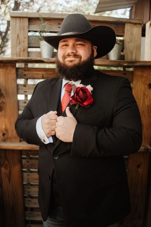 Bearded groom in black suit, red tie, boutonniere, and cowboy hat smiling at outdoor rustic wedding venue.
