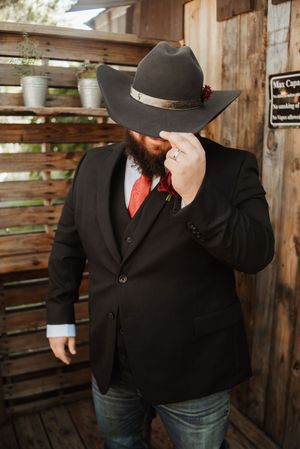 Bearded groom in black suit, red tie, and cowboy hat tips his hat while wearing a boutonniere at a rustic wedding venue.