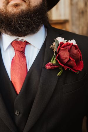 Bearded groom in black suit with red paisley tie and deep red rose boutonniere on lapel.