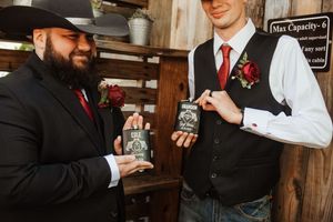 Two groomsmen in black suits with red boutonnieres holding decorative gift boxes at a rustic wedding venue.