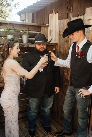 Three people in western attire toasting with shot glasses outside a rustic wooden barn at a country wedding.
