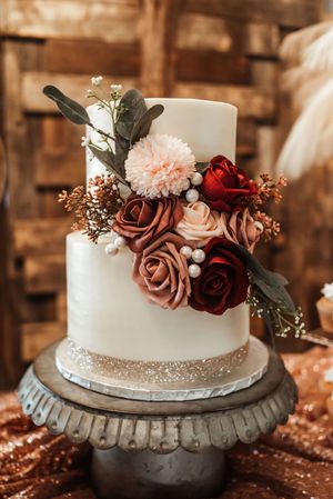 Two-tier white wedding cake with red, blush, and mauve roses, pearl accents, and gold glitter base on rustic stand.