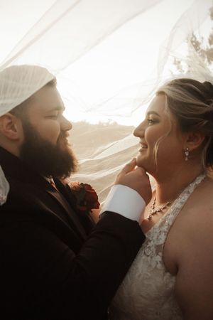 Bride and groom share a romantic moment under a flowing veil, bathed in golden light, as groom gently holds bride's chin.