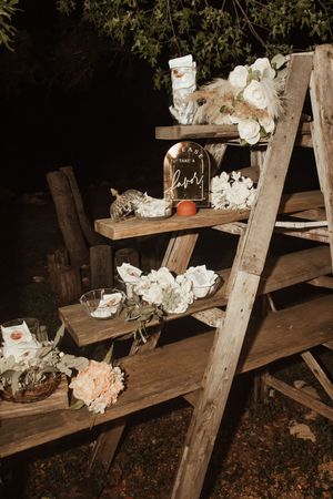 Rustic wooden ladder shelf decorated with white and peach flowers, candles, and a favor sign for an outdoor wedding reception.