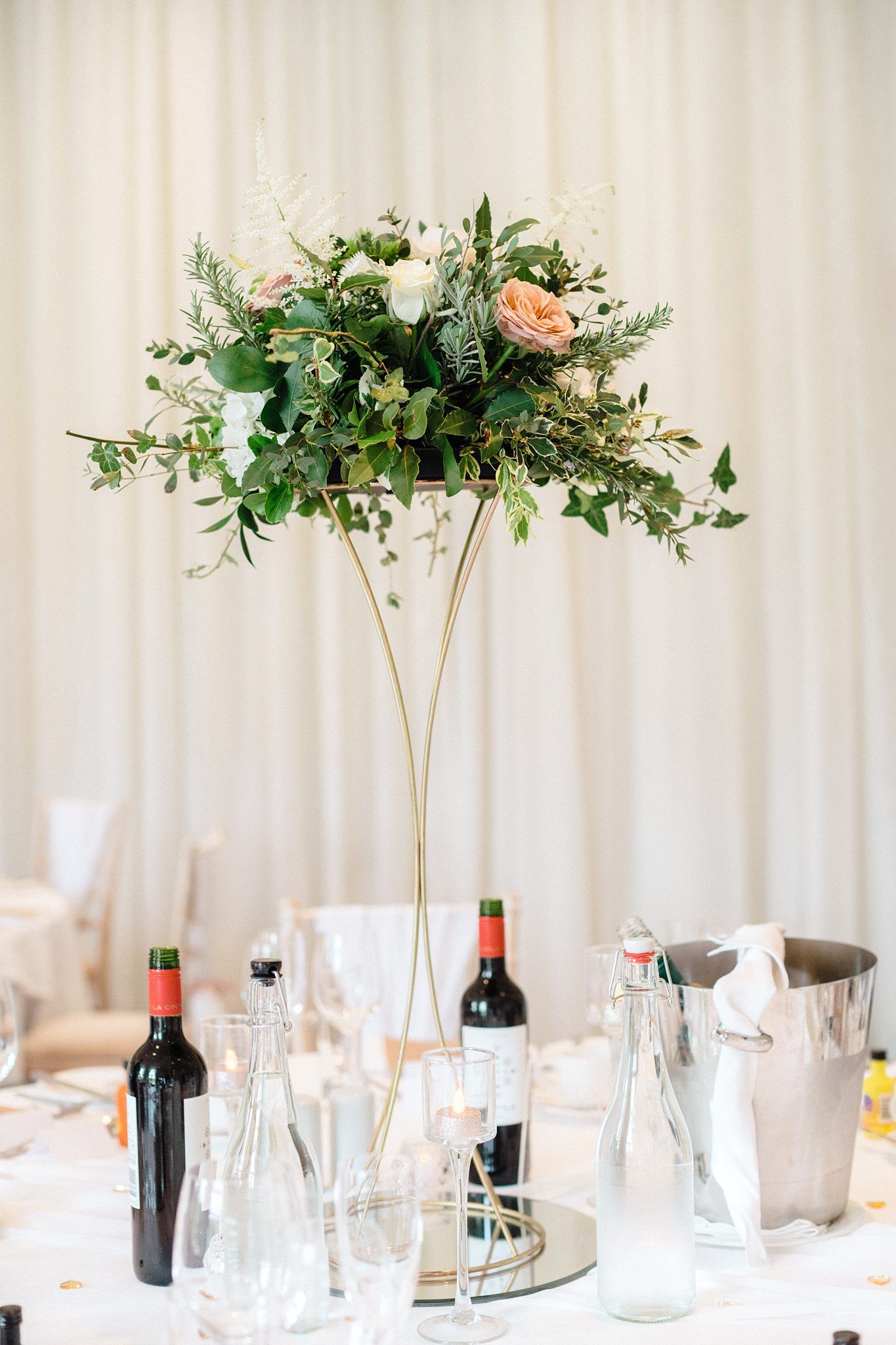 Tall gold floral centerpiece with pink roses and greenery on a wedding reception table with wine bottles and glasses at The Prince George