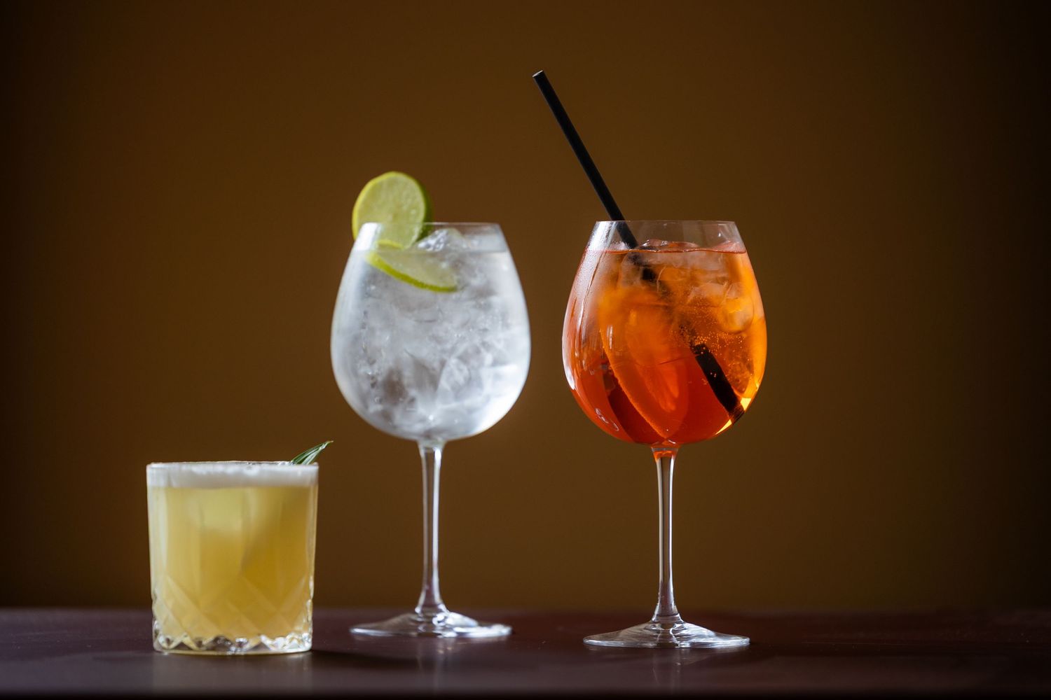 Three cocktails on a bar: a small yellow drink, a large gin and tonic with lime, and an Aperol Spritz with a straw.
