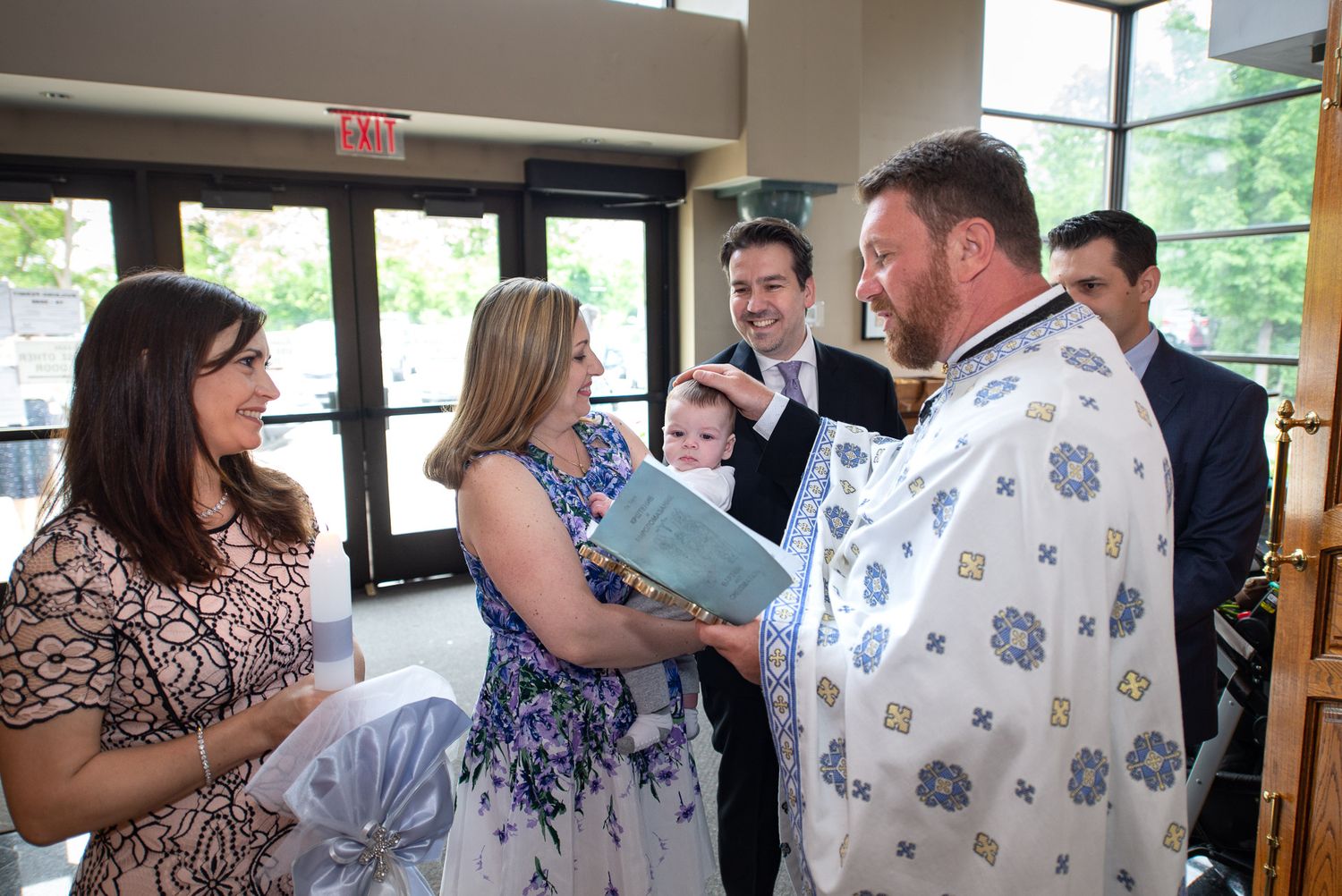 Orthodox priest in embroidered vestments greeting family with baby at baptism ceremony.