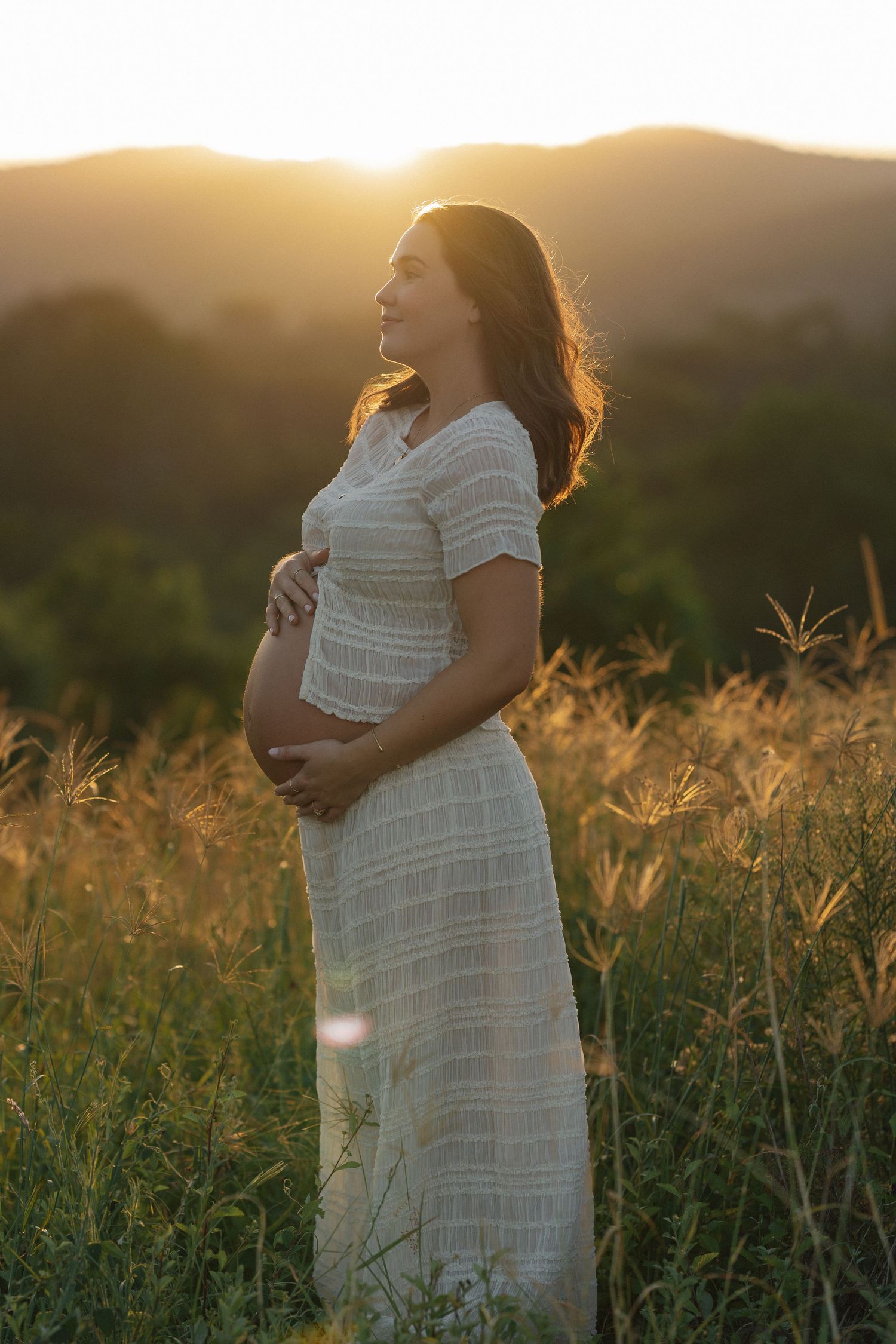 Pregnant woman in white dress cradling her baby bump in a golden sunset field with mountains in the background.