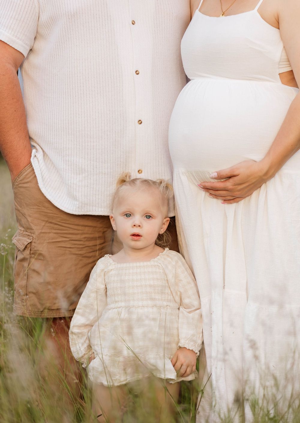 A pregnant woman in white dress with toddler girl and man in neutral tones posing in a green field.