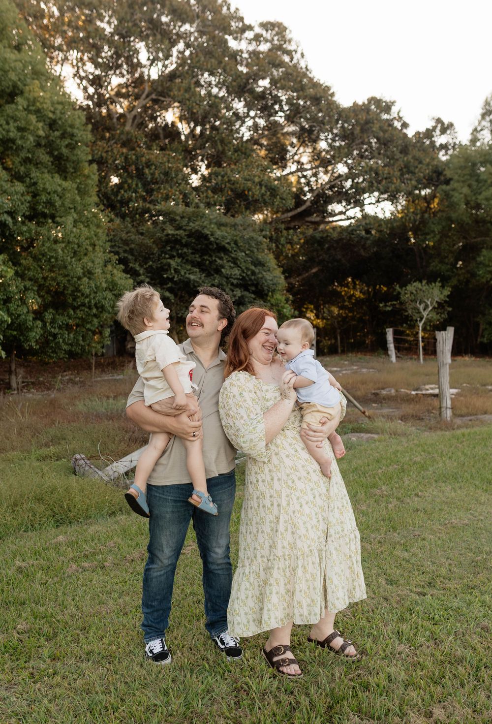 A happy family of four poses outdoors at golden hour, parents laughing while holding two young toddlers in a lush green park.